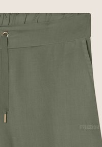 Pantalones verde oliva con cintura fruncida, cierre de cordón y detalles en dorado. Presentan una marca bordada a lo largo del costado.