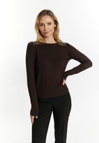 Brązowy sweter z długim rękawem o prążkowanej fakturze i ozdobnych ramionach. W połączeniu z czarnymi spodniami, modelka stoi z ręką na biodrze na białym tle.