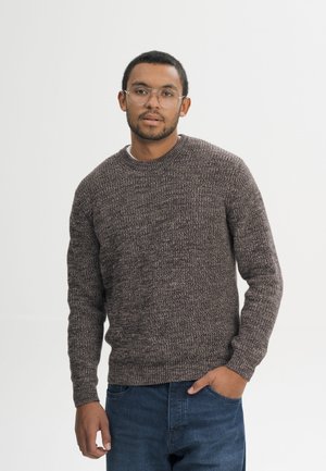 Junger Mann mit Brille, der einen braunen Strickpullover und blaue Jeans trägt, steht mit einer Hand in der Tasche vor einem einfarbigen Hintergrund.