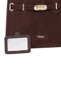 Brown suede handbag with a gold-tone clasp and a matching luggage tag. The tag features a clear window for identification. Dune London branding visible.