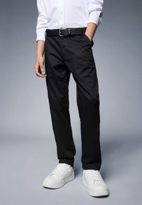 Persona che indossa pantaloni neri, camicia bianca con bottoni, cintura nera e sneakers bianche, in piedi contro uno sfondo grigio uniforme.