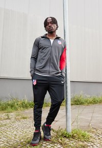 Sport tracksuit i grå og rød, der har Atletico Madrid-logoet. Sorte bukser, sorte sneakers med røde detaljer, stående nær en stang.