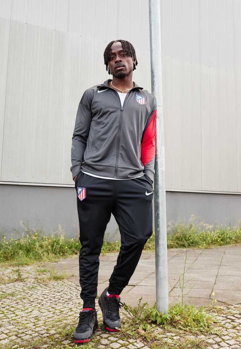 Track de sport en gris et rouge, arborant le logo de l'Atletico Madrid. Pantalons noirs, baskets noires avec des accents rouges, se tenant près d'un poteau.