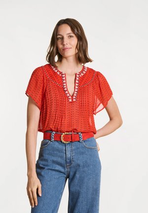 Femme portant un chemisier rouge à motifs avec un col brodé, un jean bleu taille haute et une ceinture rouge avec une boucle tressée dorée.