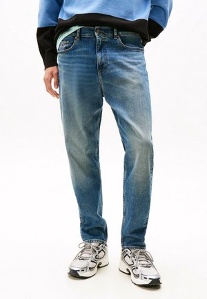 Man draagt versleten blauwe jeans, lichtgrijze en zwarte sportschoenen, en een blauw-zwart trui met handen in de zakken.