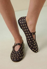 Ballerines en cuir marron avec des embellissements en clous dorés, design à lanière T, bout rond et semelle basse, mettant en avant une texture lisse et un détail discret.