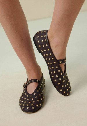 Brown leather flats with gold stud embellishments, T-strap design, rounded toe, and low sole, showcasing a smooth texture and understated detailing.