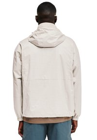 Helle beige Kapuzenjacke mit elastischen Bündchen und Saum. Das Material wirkt glatt mit einer subtilen Textur und hat eine Naht in der Mitte.