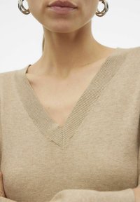 Bež pulover z V-izrezom, izdelan iz mehkega materiala, z rebrastimi poudarki na izrezu in gladko teksturo. Poglede od blizu.