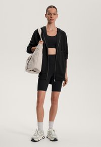 Črna jopica s kapuco na zadrgo, kombinirana s črnim športnim nedrčkom in kolesarskimi hlačami. Svetla ohlapna torba in masivni supergi. Enostaven dizajn.