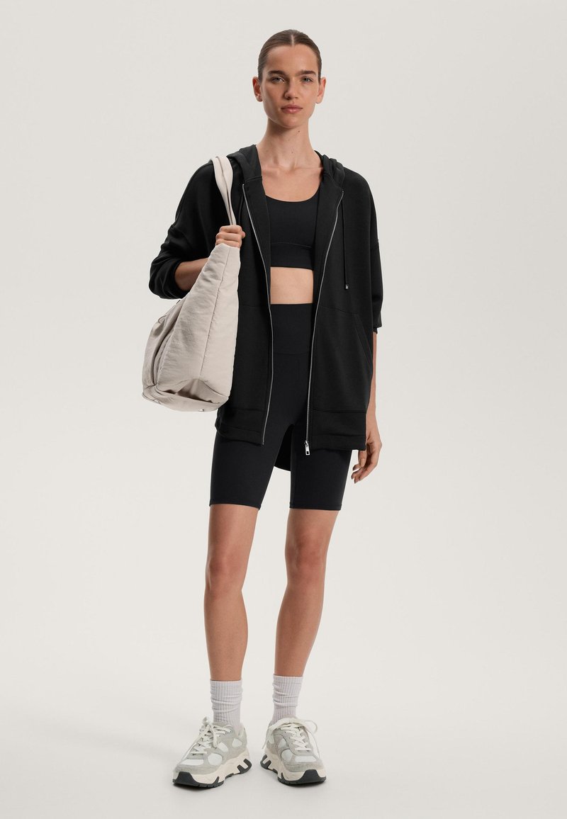 Črna jopica s kapuco na zadrgo, kombinirana s črnim športnim nedrčkom in kolesarskimi hlačami. Svetla ohlapna torba in masivni supergi. Enostaven dizajn.