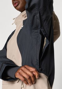 Črna in bež kapuca s podrobnostmi na zadrgo na rokavu, izdelana iz lahkega materiala z gladko teksturo in zasnovo z blokiranjem barv.