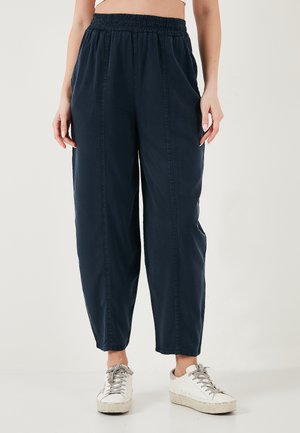 Naine, kes kannab lahtisi tumesiniseid pükse elastse värvliga ja valgeid tossusid, seisab tavalise hele tausta ees.