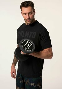Černé tričko s potiskem "UNLMTO", držící černou gumovou činku (12,5 kg) ve vzorovaných modro-zelených kalhotách, ukazující tetování na paži.