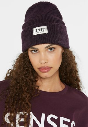 Purple knitted beanie with a folded cuff and a white label reading "SENSES THE LABEL." Background is minimal, focusing on the hat's texture.