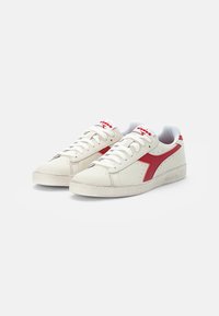 White leather sneakers with red accents and logo, featuring a textured rubber sole, perforations on the toe, and flat laces.