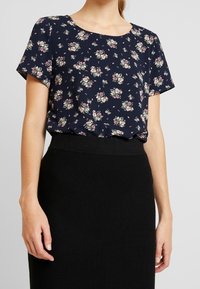 Marineblaue Bluse mit kurzen Ärmeln, verziert mit kleinen rosa und weißen Blumen, runder Ausschnitt und leichtem Stoff, kombiniert mit einem schwarzen Rock.