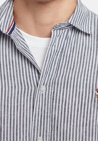 Mann in einem blau-weißen, vertikal gestreiften Hemd mit Knöpfen über einem weißen Rundhalsunterhemd, das den Kragen und den oberen Brustbereich zeigt.