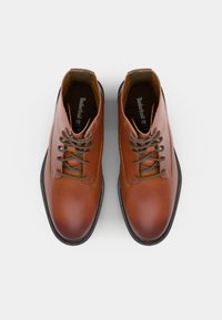 Botas de tobillo de cuero marrón con punta redonda, cordones planos y ojales grises en contraste. Suela de goma negra texturizada. Logo de Timberland visible en el interior.