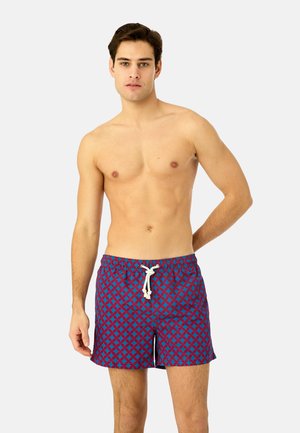 Junger Mann stehend, trägt blaue und rote gemusterte Badeshorts mit weißem Kordelzug, neutraler Hintergrund.