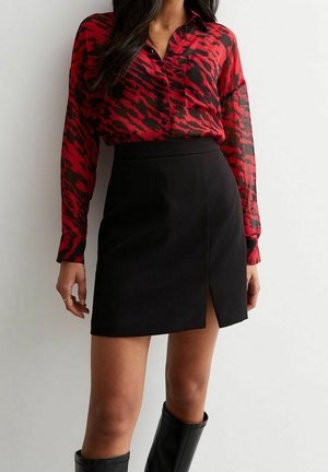 Femme portant un chemisier boutonné rouge et noir à motifs, une mini-jupe noire fendue à l'avant et des bottes noires montantes jusqu'aux genoux.