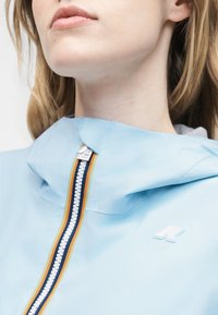 Κοντινό πλάνο ενός ατόμου που φοράει ένα γαλάζιο μπουφάν με φερμουάρ με πολύχρωμες ρίγες και ένα μικρό λευκό λογότυπο στο στήθος.