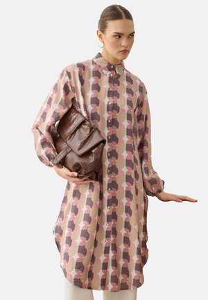 Vrouw met een lange, patroonknopen blouse die een grote bruine, getextureerde clutch vasthoudt tegen een effen achtergrond.