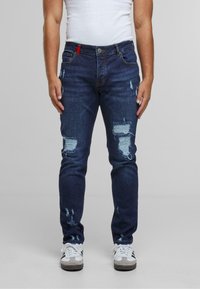 Mørkeblå denim jeans med slitte hull, slim fit, fem-lomme design, og en rød aksent på livet. Kombinert med hvite joggesko.