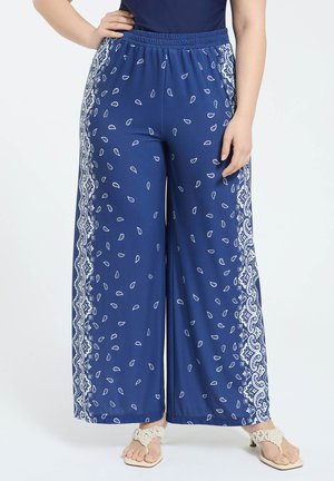 Femme portant un pantalon bleu à jambes larges avec des motifs paisley et géométriques blancs sur les côtés, associé à des sandales à talons beiges.