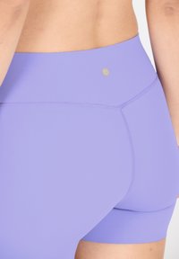 Nahaufnahme einer Person, die hoch taillierte lila Sportshorts mit einem kleinen goldenen runden Logo auf dem hinteren Bund trägt.