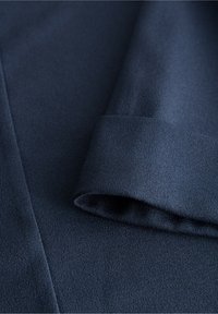 Tmavě modrá texturovaná látka s hladkým povrchem a detailem v podobě zatočeného lemu. Látka působí měkce a jemně se zavěšuje.