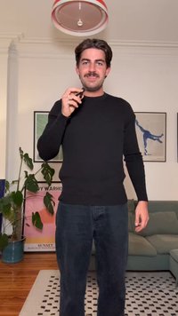 Svart långärmad tröja med figursydd design, kombinerad med mörka byxor. Bakgrunden har växter och färgglada inramade konstverk.