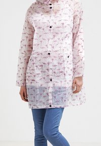 Transparent regnjacka med mönster av rosa flamingos, tryckknappar, en huva och två framfickor. Ärmarna är långa och lösa.