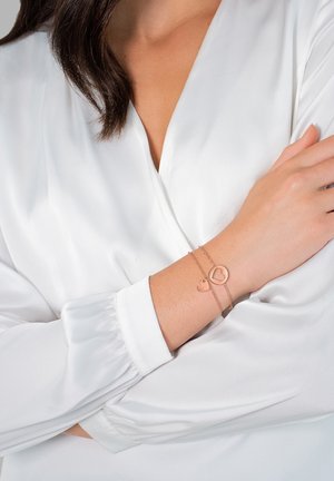 Bracelet en or rose avec un charm rond et un charm en forme de cœur, porté sur un poignet contre une blouse blanche au toucher doux et lisse.