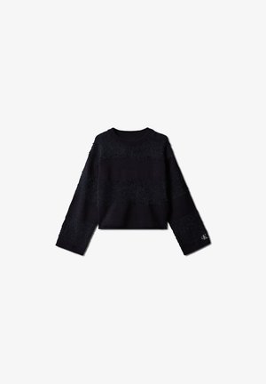 Junges Mädchen mit langen Haaren, das einen schwarzen Pullover, dunkle, weite Jeans und schwarze Loafers trägt, steht mit den Händen in den Taschen.