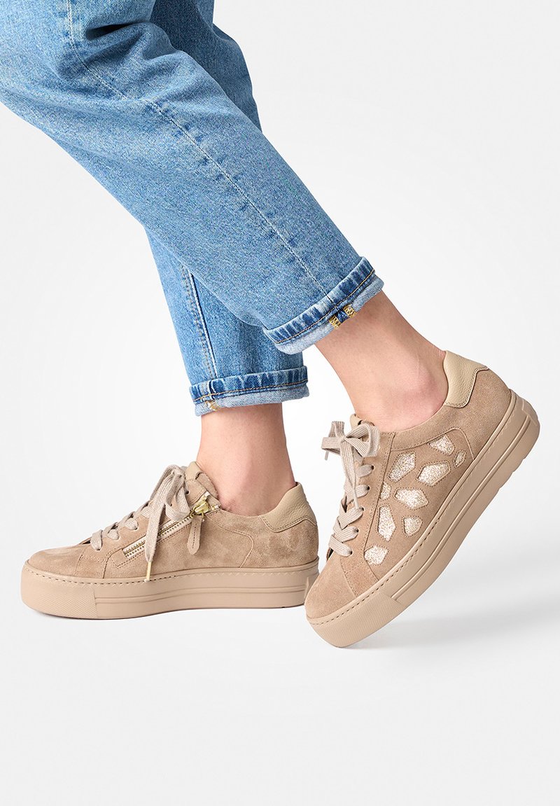 Tan suede platform sneakers with side zipper and glittery cutout details worn with rolled-up blue jeans on a white background.