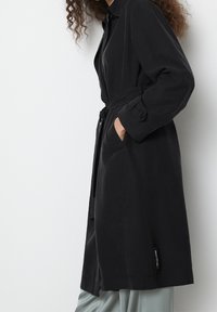 Černý trenčkot s páskem v pase, dlouhými rukávy a manžetami na knoflíky. Má hladkou texturu a cedulku poblíž lemu.