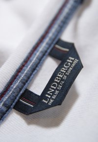 Blaues Textilkennzeichen mit weißen und roten Streifen, das den Text "LINDERGH DAS BLAUE GARANTIE-SIEGEL" auf einem weißen Stoffhintergrund zeigt.