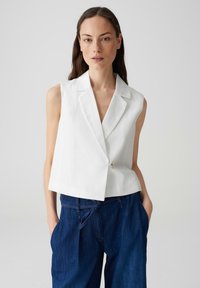 Blazer bianco senza maniche con colletto a punte e chiusura a un bottone, abbinato a jeans larghi blu scuro. Texture liscia, design minimale.