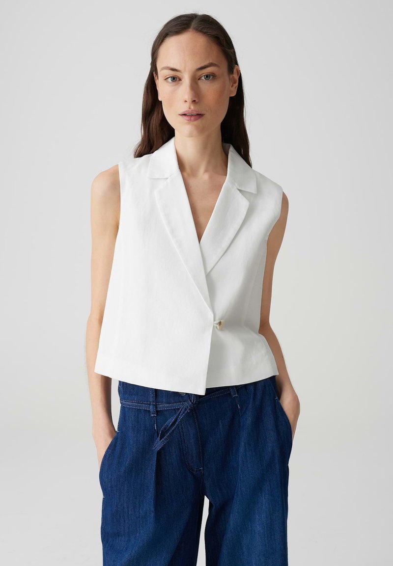 Blazer bianco senza maniche con colletto a punte e chiusura a un bottone, abbinato a jeans larghi blu scuro. Texture liscia, design minimale.