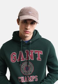 Gorra de pana marrón con logotipo bordado en la parte delantera, combinada con una sudadera verde con el gran texto "GANT" y un gráfico circular.