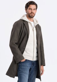 Manteau en laine vert foncé au design ajusté, avec deux boutons, un col cranté, superposé à un sweat à capuche zippé gris clair.
