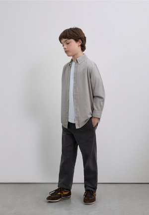 Camisa de botones gris claro sobre una camiseta blanca, combinada con pantalones de pana oscuros y zapatos marrones con detalles amarillos. Diseño sencillo, ajuste relajado.