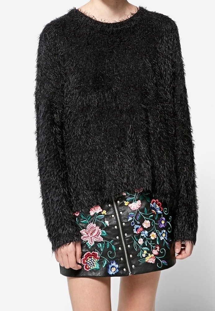 Pull noir duveteux à manches longues associé à une mini-jupe en cuir noire ornée de broderies florales colorées et d'une fermeture éclair à l'avant.