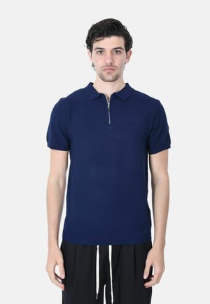 Uomo che indossa una polo a maniche corte blu navy con una cerniera frontale, abbinata a pantaloni neri con cordini bianchi.