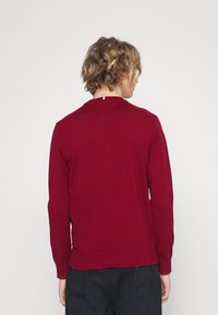 Suéter vermelho de mangas longas com um decote redondo, apresentando uma faixa de cor horizontal na parte de trás do colarinho e uma bainha canelada.