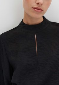 Primo piano di una persona che indossa una maglia nera texturizzata con un alto colletto e un'apertura a chiave verticale al centro davanti.