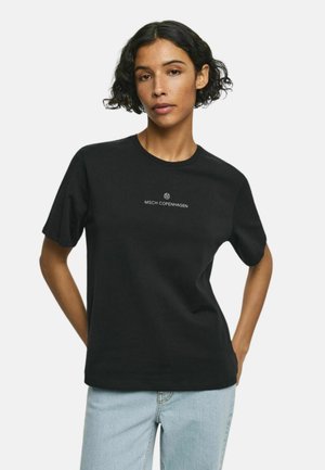 Kobieta o krótkich kręconych włosach, ubrana w czarną koszulkę MSCH Copenhagen i jasnoniebieskie dżinsy, pozująca z rękami za plecami.