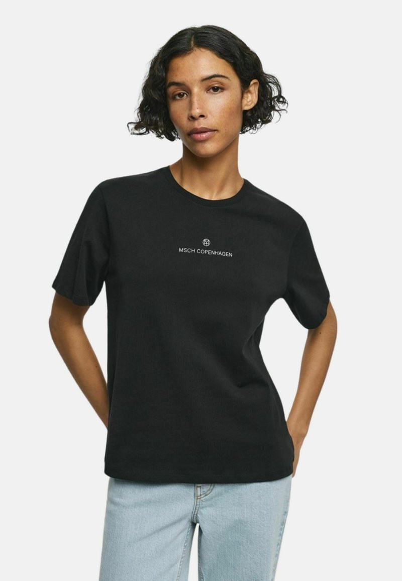 Žena s krátkými kudrnatými vlasy, oblečená v černém tričku MSCH Copenhagen a světle modrých džínách, pózující s rukama za zády.