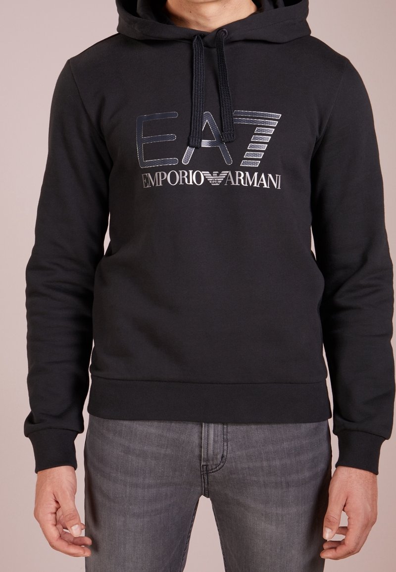 Homme portant un sweat à capuche noir EA7 Emporio Armani avec cordons de serrage et jean gris, debout devant un fond uni.